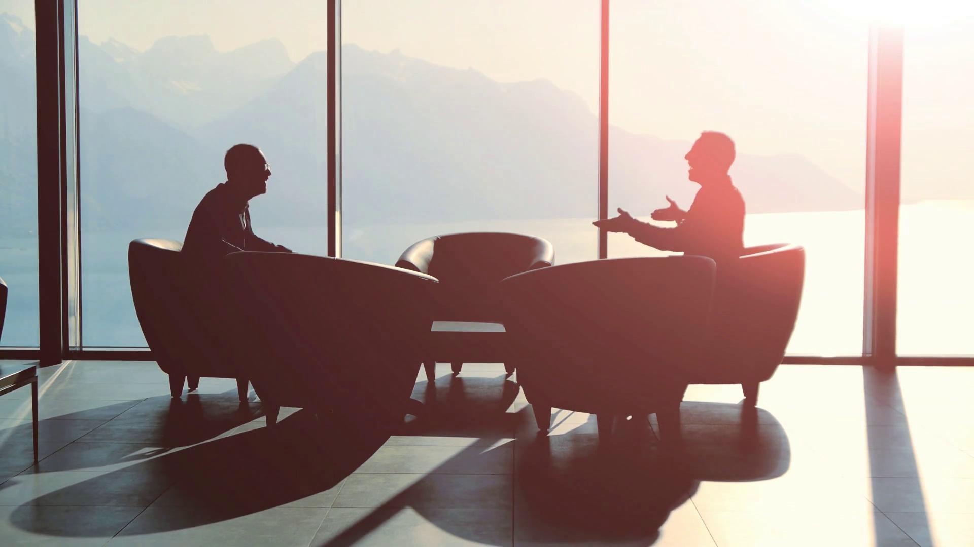 Two people in conversation in a glass-walled room above a mountain lake.
