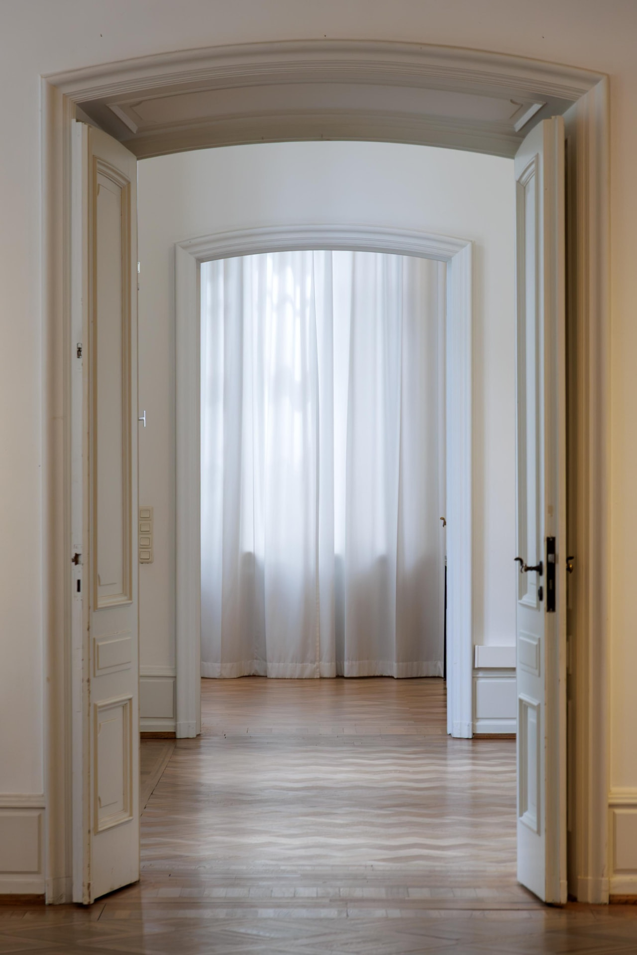 An arched enfilade of a historic apartment with parquet floors and sheer curtains.