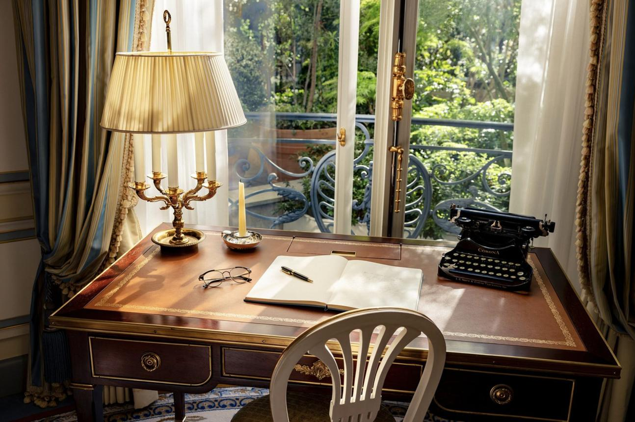 A writing desk with an open notebook, fountain pen and typewriter beside open balcony doors.